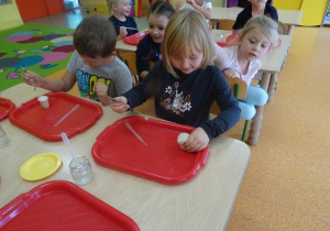 Dzieci siedzi przy stoliku, obserwują reakcje wody utlenionej z drożdżami w swoich pojemnikach. Mieszają składniki za pomocą łyżeczki.